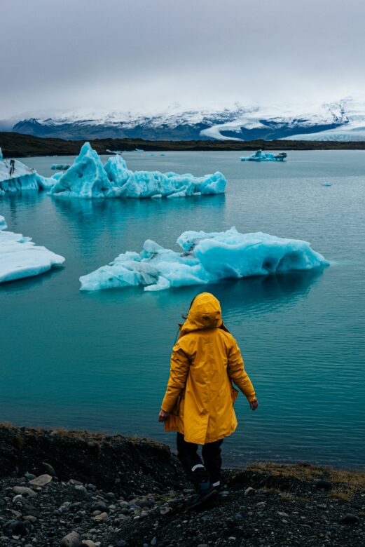 A person is wearing their poncho standing on the edge of a large lake with ice glaciers.
